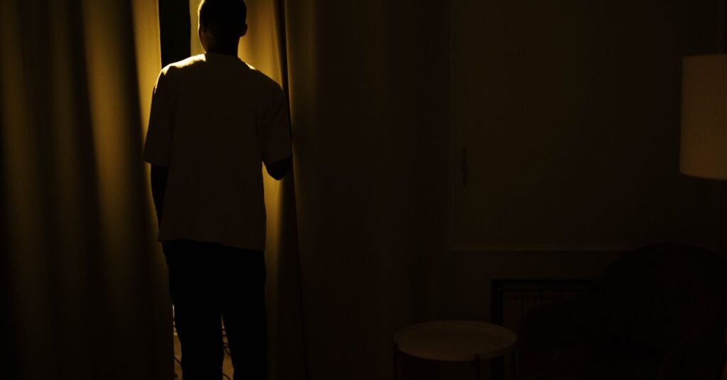 夜明けの光が部屋を満たすなか、窓のそばに立つ男性。こわばっていた肩が、ゆっくりとほどけていく静かな変化。
