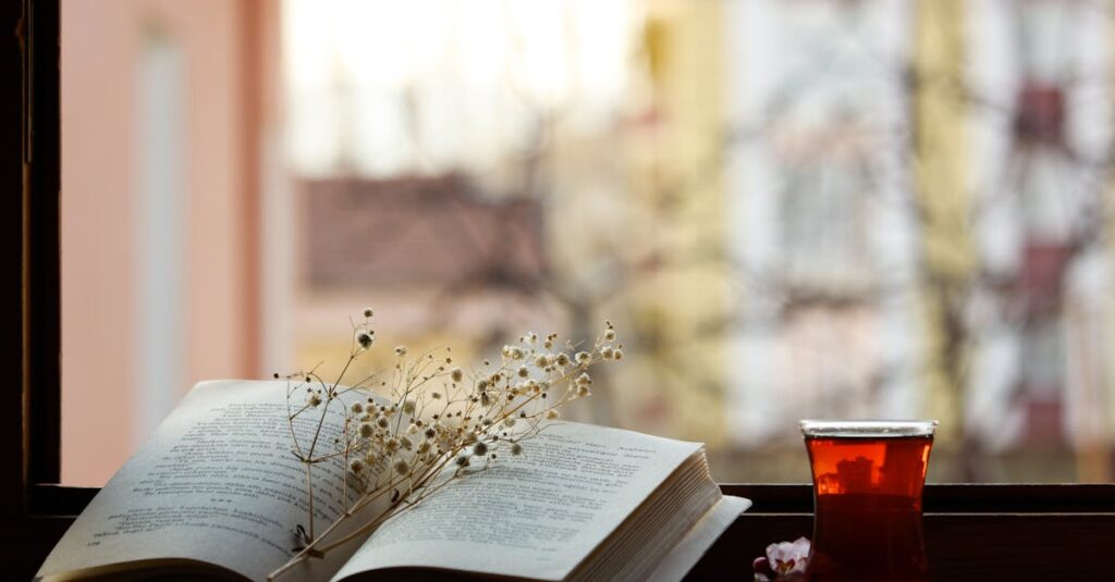 窓辺に置かれた開かれた本とお茶。ろうそくの光があたたかく揺れる、静かな読書の情景。