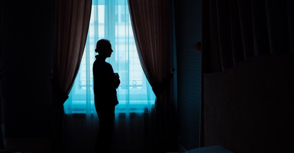 夜明けの光が部屋を満たすなか、窓のそばに立つ男性。こわばっていた肩が、ゆっくりとほどけていく静かな変化。
