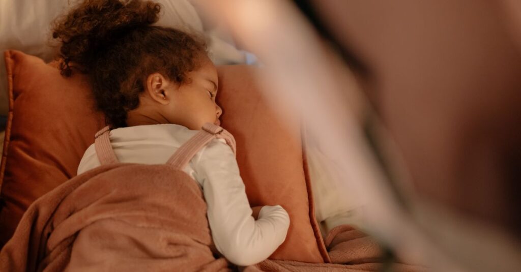 暖炉のそばの椅子で毛布にくるまって眠る子供。誰かがそっとかけてくれたような、安心のぬくもり。